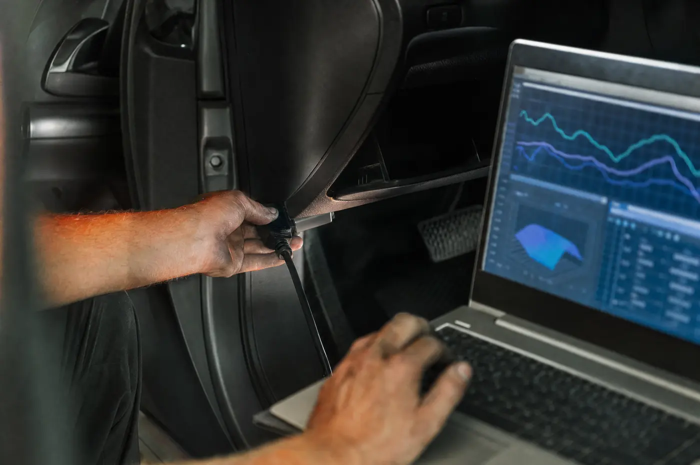 Mekaanikon kädet liittämässä diagnostiikkakaapelia auton OBD-liittimeen, kannettavan näytöllä käyriä